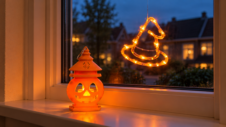 PumpkinGlow-lantaarn op een vensterbank met een PumpkoGlow-heksenhoed als lichtfiguur aan het raam